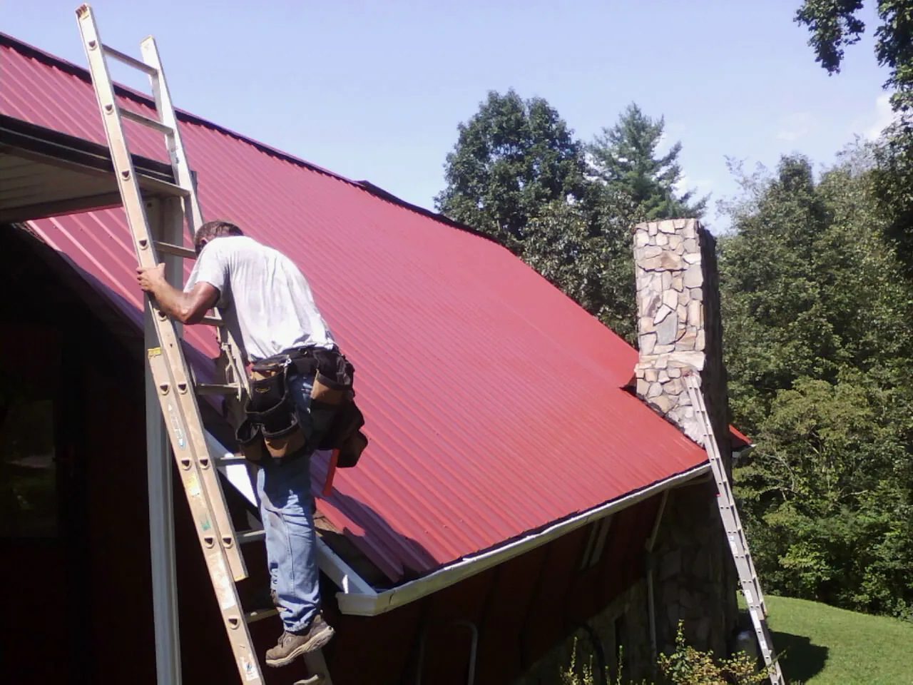 Roofer on a ladder beside a red metal roof and stone chimney — Soffit & Fascia Repair by Roofing Brothers in East Palestine